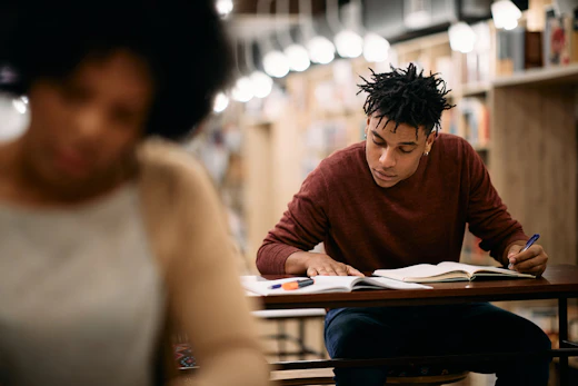 A student focused on writing notes from a book in the quiet environment of a library. A male student seated at a library desk writing in a notebook, with bookshelves and blurred study lights in the background.